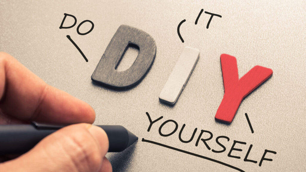 Hand holding a pen writes "YOURSELF" under 3D letters "DIY" on a textured surface. Words "DO IT" encircle "DIY," conveying a motivational tone.