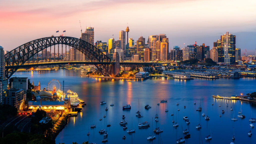 Sydney Harbour Bridge and city skyline silhouetted against a vibrant sunset sky, casting warm hues over the water.