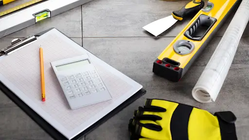 Construction planning tools including clipboard, calculator, gloves, measuring level, and blueprints on a tiled surface.
