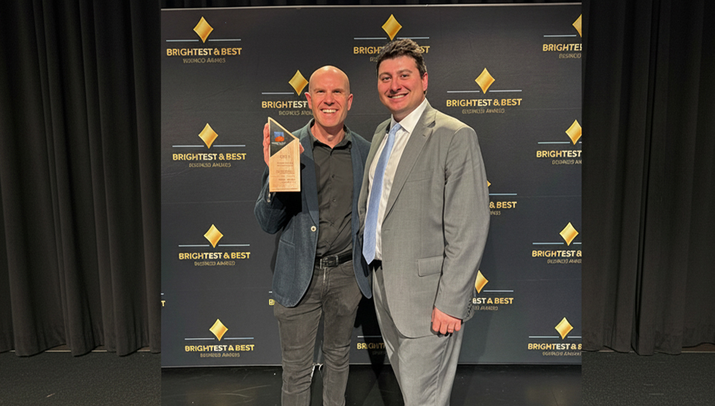 Two men pose together at the Brightest & Best Business Awards; one is holding a wooden and glass trophy while standing in front of a branded step-and-repeat backdrop.